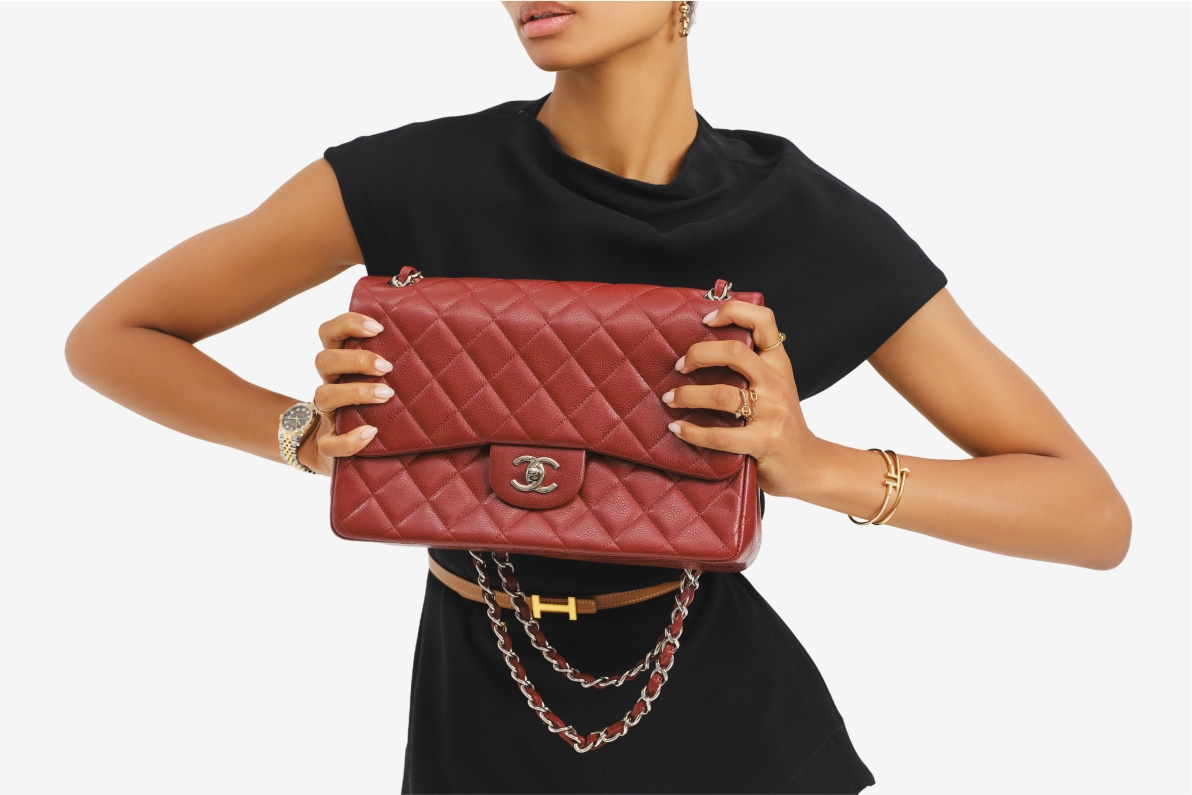a woman wearing a short sleeve black dress and a tan Hermes belt with a yellow gold H buckle and holding a burgundy Chanel Jumbo Double Flap bag