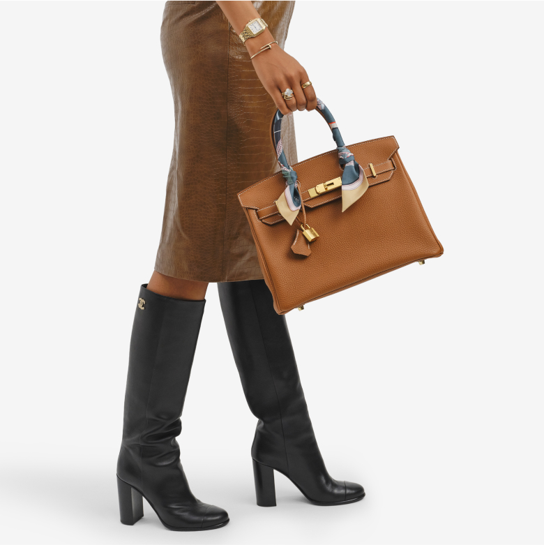a woman wearing a brown leather midi skirt and black Chanel knee high boots and holding a gold color Hermes Birkin 30 bag